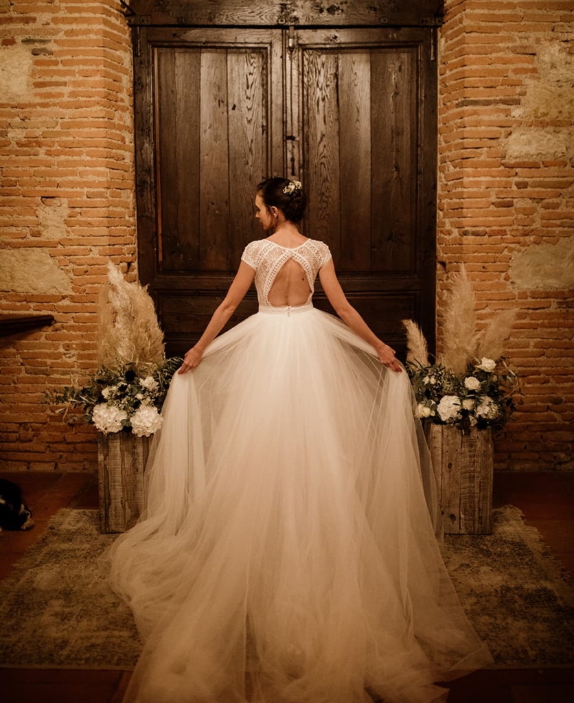 Mariée en robe blanche devant une porte en bois ancienne flanquée d'arrangements floraux blancs champêtres