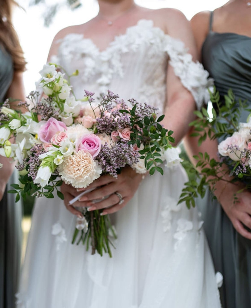 Bride and bridesmaids holding romantic bouquets with blush roses, white flowers, and dusty purple accents