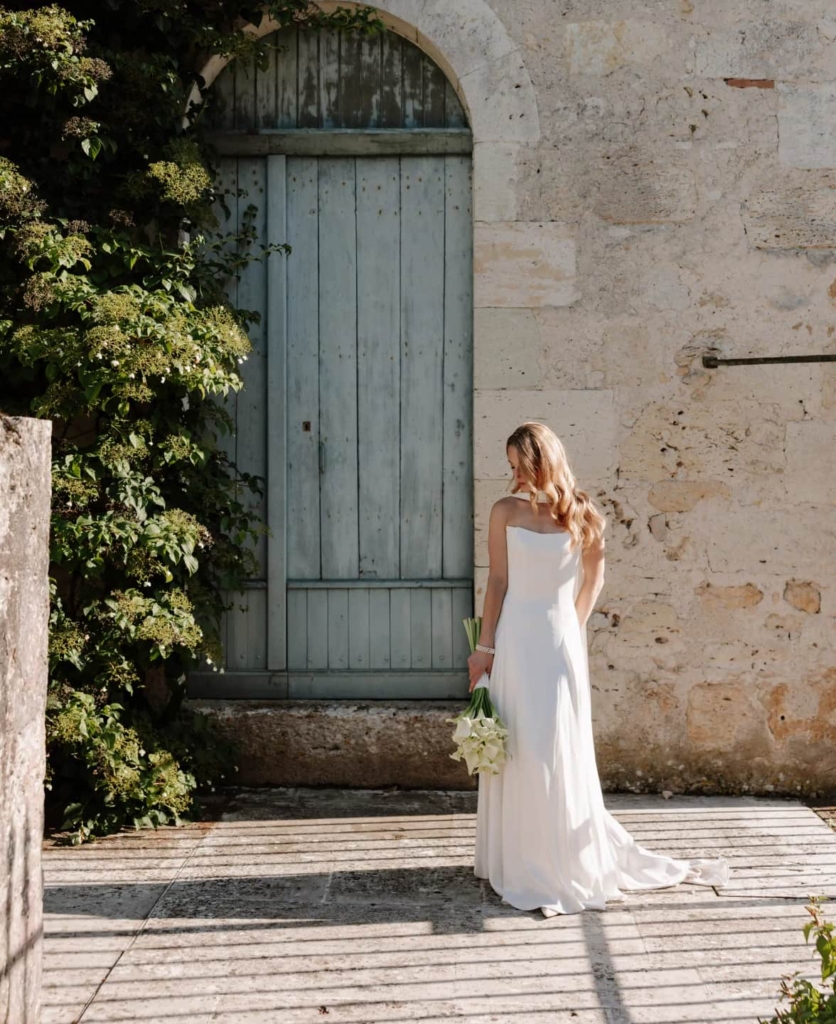 Bride in white gown holding white bouquet against weathered stone chateau wall and arched door