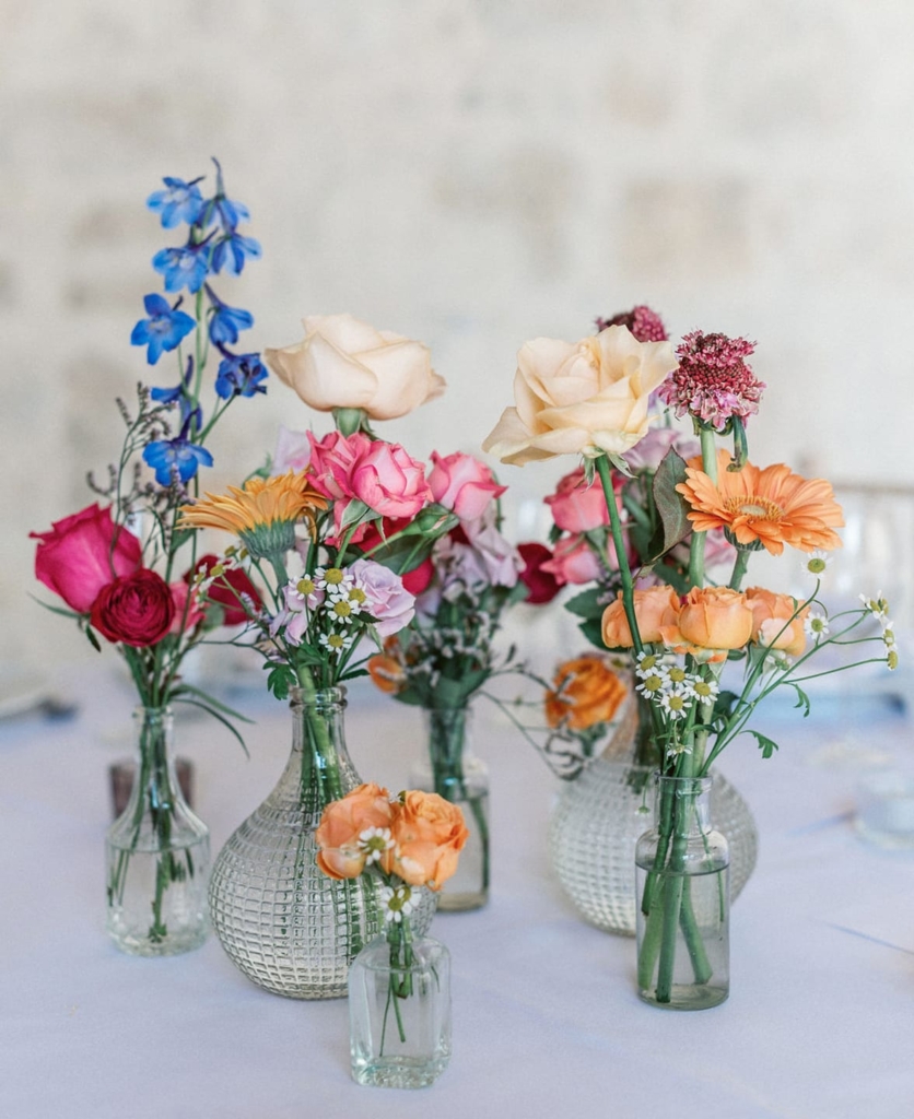Collection of seasonal wedding flowers in individual glass vases including roses, delphiniums, gerberas, and astilbe arrangements