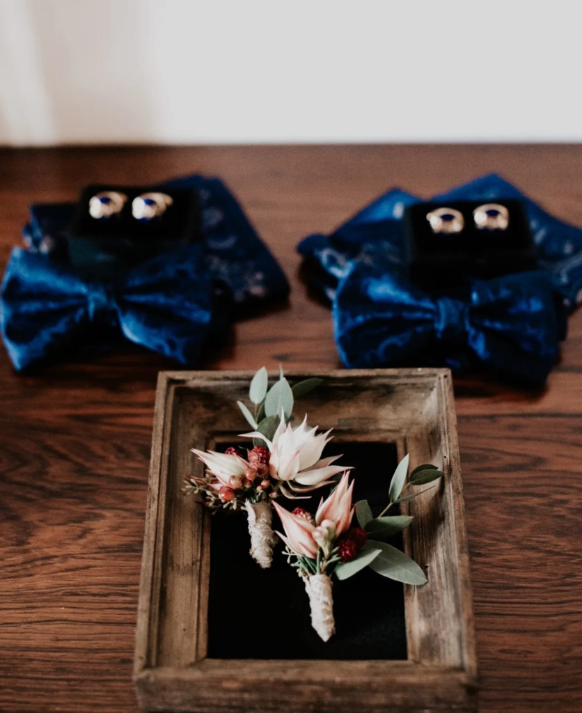 Boutonnières florales blanches et rouges avec eucalyptus dans cadre bois rustique, nœuds bleus à l'arrière