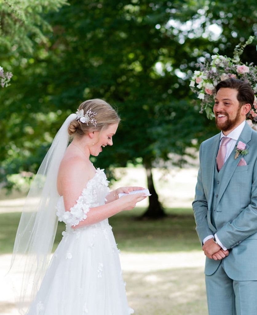 Mariée en robe blanche et voile sous arche fleurie rose et blanc avec marié en costume bleu