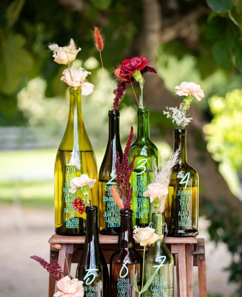 Bouteilles de vin gravées avec noms de convives, habillées de fleurs roses, blanches et amarante sur support en bois