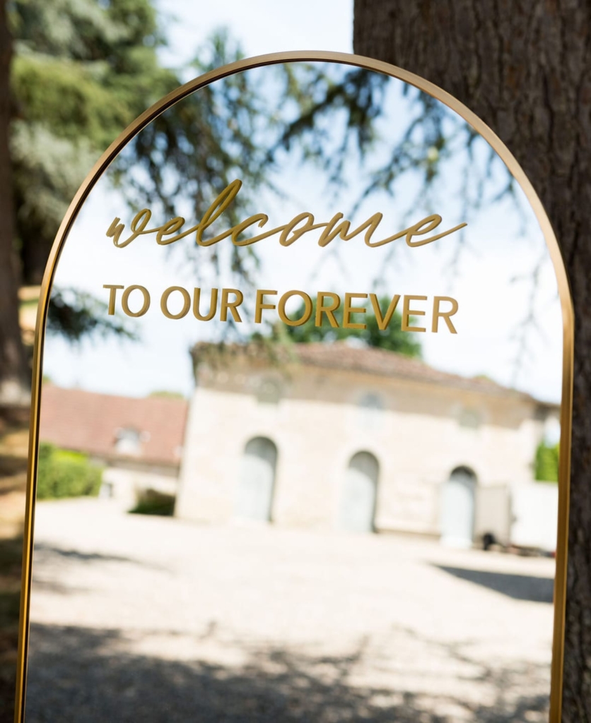 Gold arch mirror with welcome to our forever text at outdoor wedding venue entrance